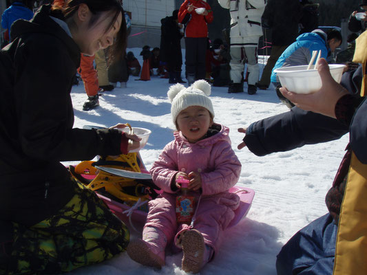 白い帽子をかぶり、ピンク色のスキーウエアを着てピンク色のそりに座っている女の子と横でキノコ汁を食べさせようとしている母親の写真
