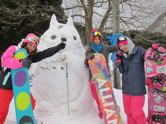 雪だるまの前でスノーボードの板をもって立っている3人の女性の写真