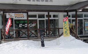 ガラス窓にヒュッテ ビーウィンドと書かれたお店とお店の前に立っているのぼり旗、積もった雪とラックに立てかけられたスノーボードの写真