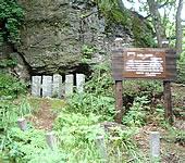 崖面の下にぽっかりと穴が開いた洞窟の右側手前に、木製の立て看板が設置されている写真