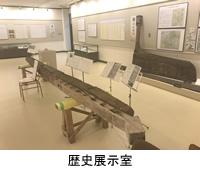 展示室の広いスペースに資料が置いてある4つの楽譜立てがあり、手前に細長い出土品が設置されている歴史展示室の写真