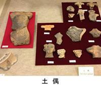 左側に縦長の赤色の敷物、右側にL字型の茶色の敷物の上に発掘された顔の形など様々な形をした土偶が並んで置いてある写真