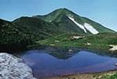 少し雪が残った緑色の山の手前に湖がある飯豊山の主稜を写した写真