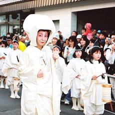 白無垢姿の女性とその横で並んで歩く、真っ白い衣装を着た稚児たちの行列を沿道でたくさんの人たちが見学している写真
